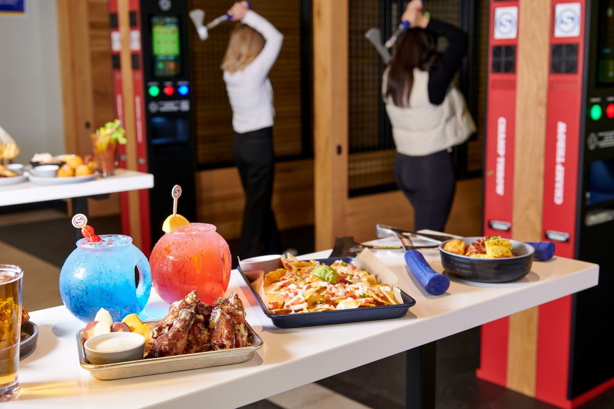 A white counter set with trays of chicken wings, nachos, and blue and pink fishbowl cocktails, with two people participating in axe-throwing behind the counter in the background.