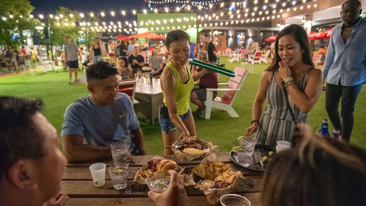 A group of people sitting at a picnic table eating burgers and other food in an outdoor space with turf grass, lit by strings of globe lights, with other people socializing in the background.