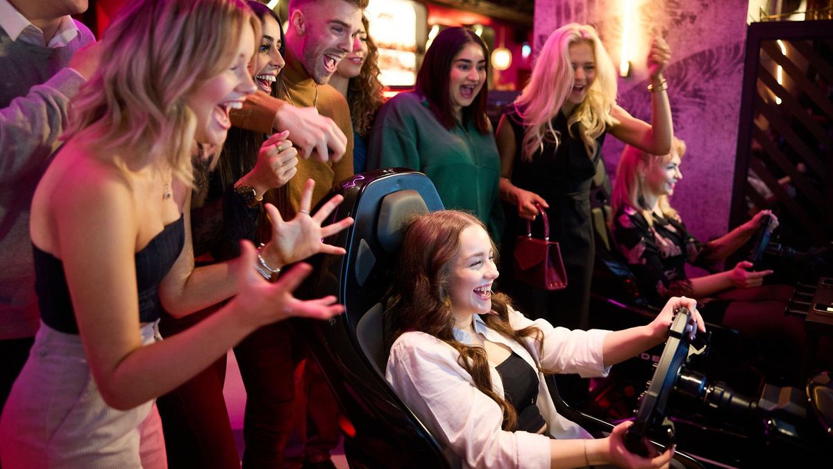A person doing simulated auto racing and smiling, with a group of friends gathered behind her cheering her on. 