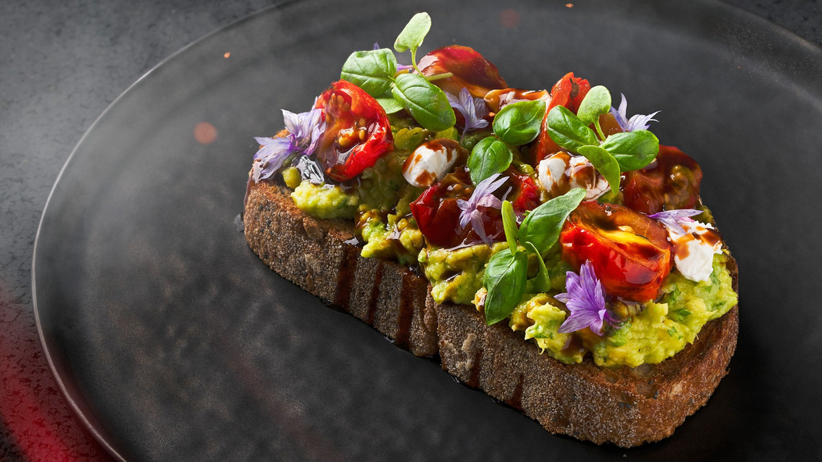 A thick slice of toast topped with smashed avocado, basil, tomatoes, balsamic vinegar, and purple edible flowers. 