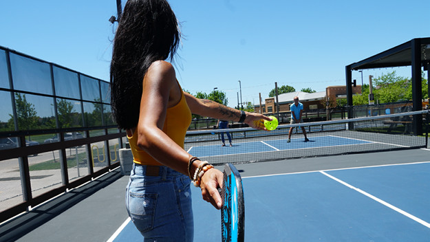 A person with long black hair and an orange shirt and jeans about to serve in a pickleball game on an outdoor court. 