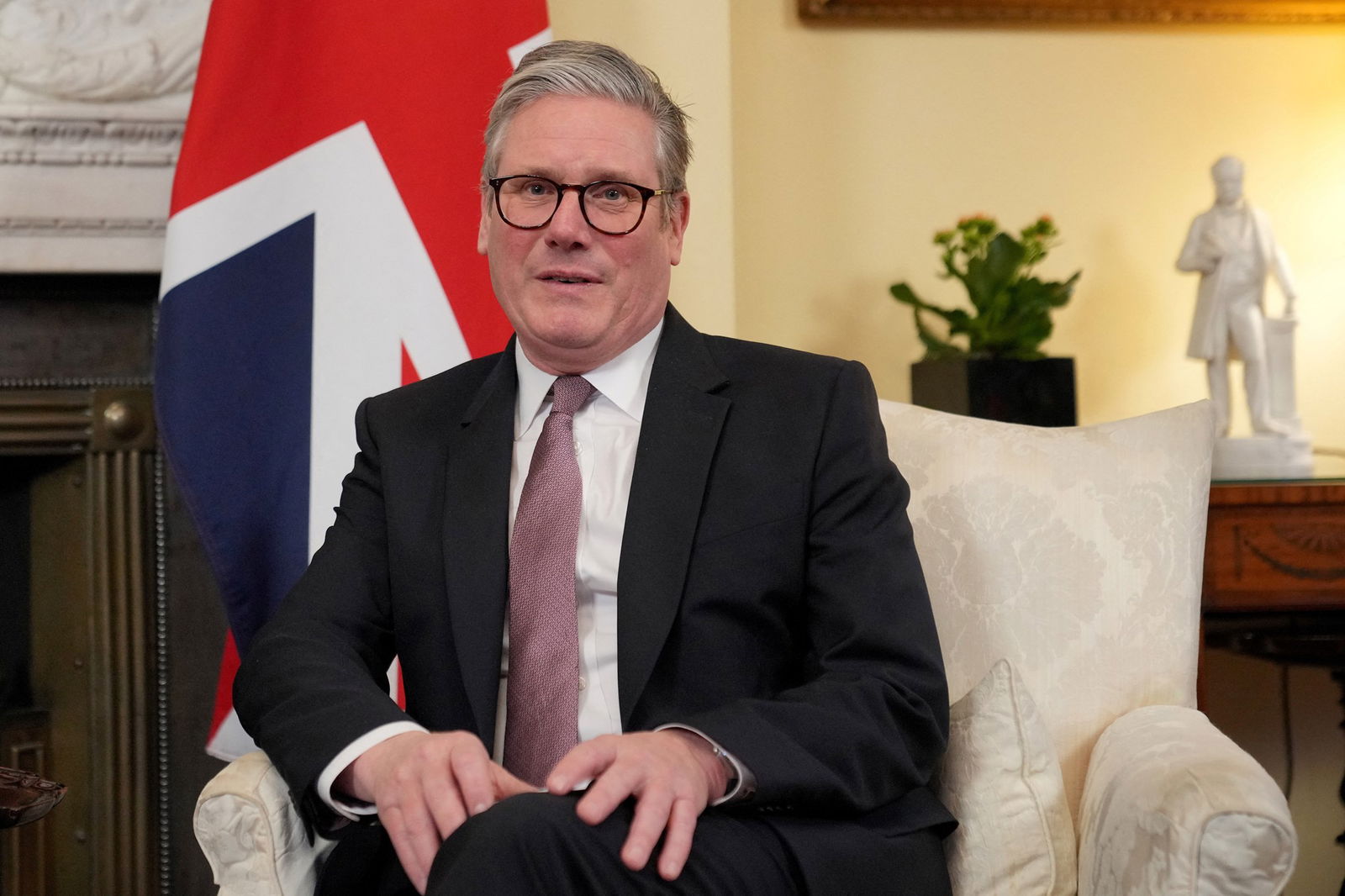 Keir Starmer sits in a white chair in front of an ornate fireplace and an English flag.