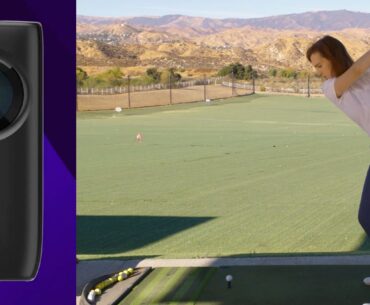 A woman in a white shirt and blue pants is preparing to hit a golf ball on a green driving range with mountains in the background. To the left, there is a close-up of a black cylindrical device on a purple background.