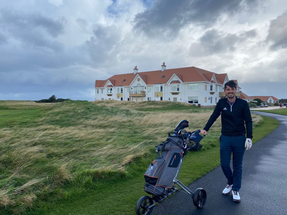 Golfer standing on a golf course with a golf cart in front of a large house.