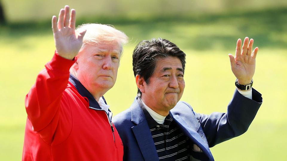 US President Donald Trump with the late Japanese Prime Minister Shinzo before playing a round of golf at Mobara Country Club in Chiba, Japan on 16 May 26, 2019. Trump is wearing a red jacket and Abe has on a dark blue blazer and blue polo shirt with white stripes. They are both waving.