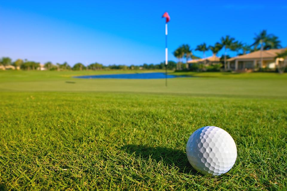 A golf ball rests near the green and pin of a tropical golf course with a clear sky above.