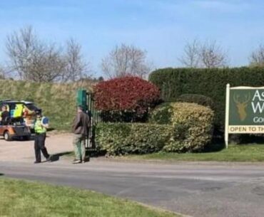 A gate on a road with green hedges on either side and a police car parked on the other side of the entrance. A police officer in a high-vis jacket and black hat can be seen at the entrance near a man with his back to us wearing a brown jacket and green trousers.