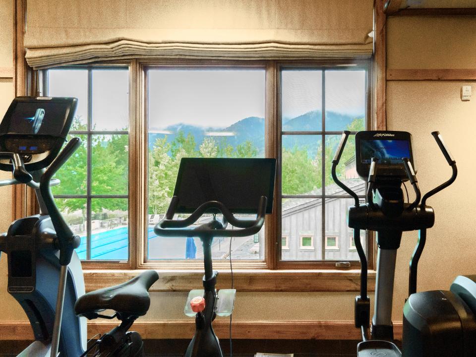 Three elliptical machints with screens face a window with a view of a pool and a barn in front of mountains on a hazy day