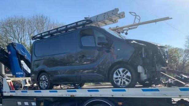 A grey van, with damage to the front of it, on the bed of a two truck.