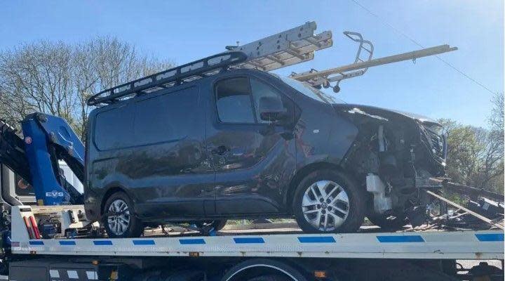 A dark grey van with damage to the front and a ladder on the roof on the back of a trailer with trees in the background
