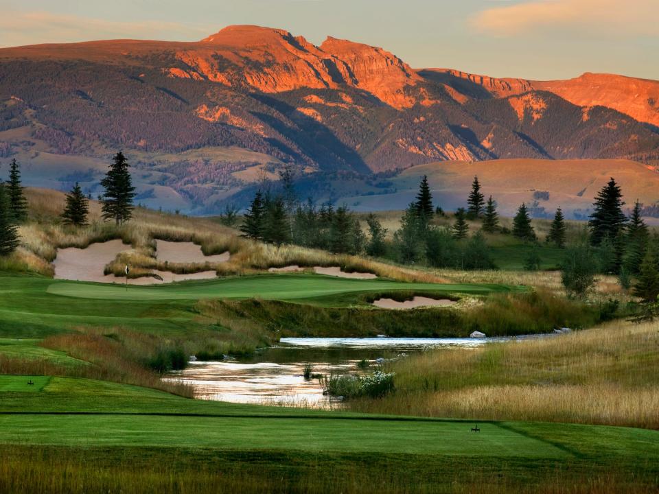 A gold course divided by a stream and dotted with Aspen trees and sand dunes sits in front of a mountain at sunset