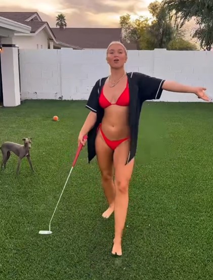 Woman in a red bikini and black shirt playing golf on a lawn with a small dog.
