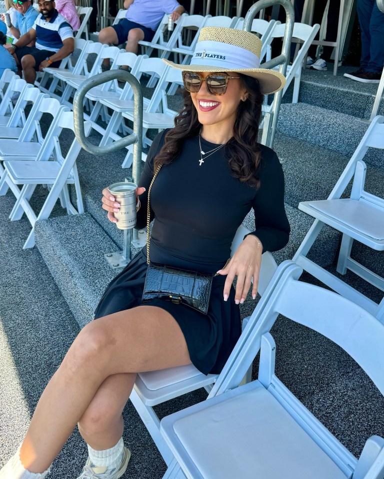Woman in a black dress and hat sits in a chair at a golf tournament, holding a drink.
