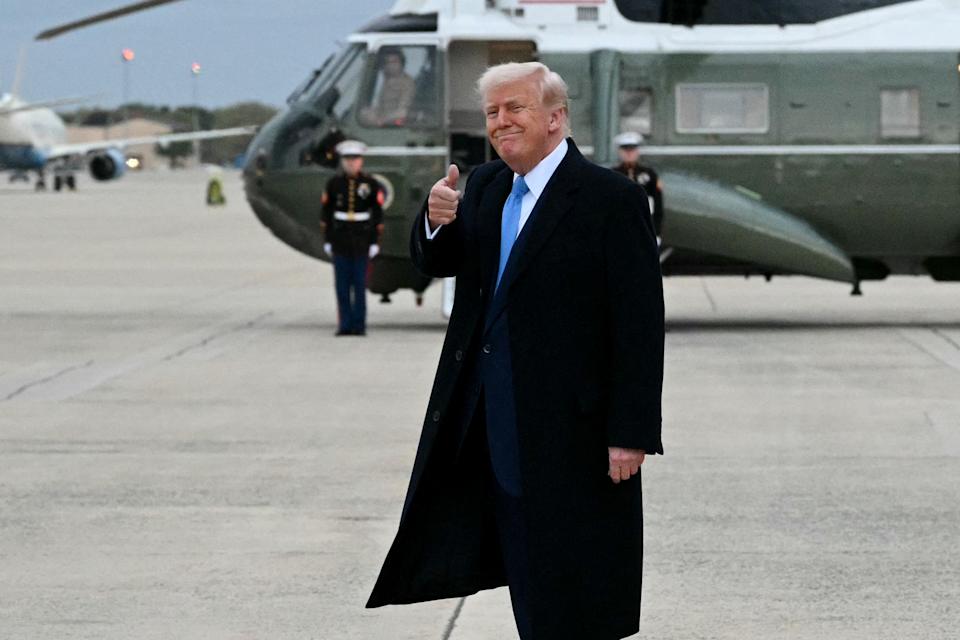 Person in a coat giving a thumbs-up gesture while standing near a helicopter on an airfield. Two military personnel are visible in the background