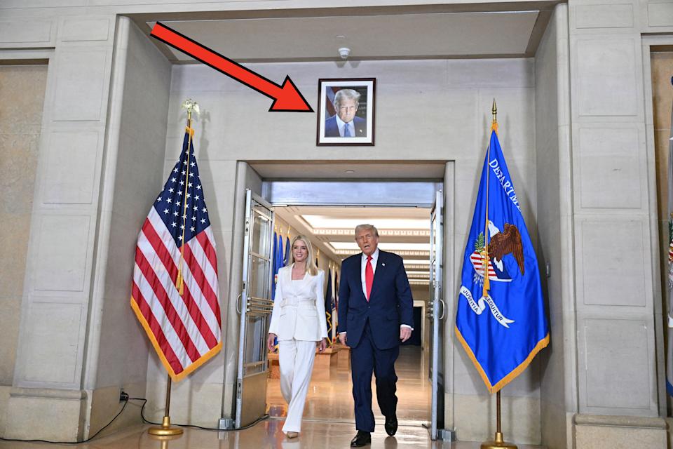 Two people in formal attire, walking indoors between U.S. and Department of Defense flags, with a framed portrait above