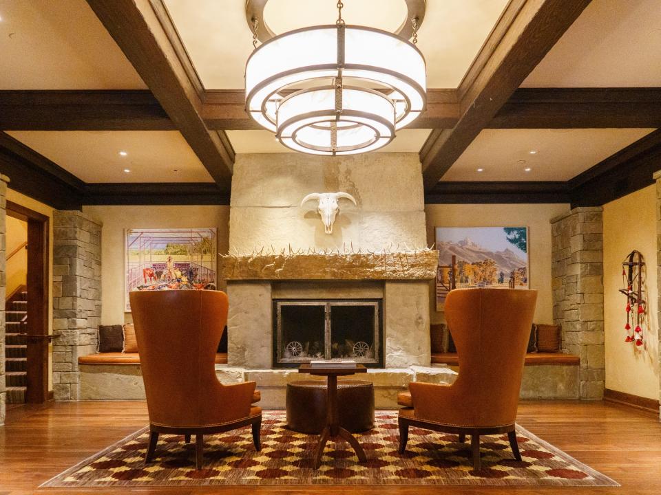 A room with stone brick pillars on the walls, two chairs and a table between them, and a fireplace in front of the chirs with an animal skull on the wall above it