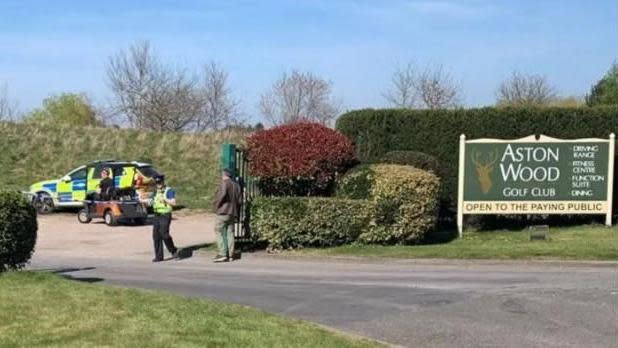 A gate on a road with green hedges on either side and a police car parked on the other side of the entrance. A police officer in a high-vis jacket and black hat can be seen at the entrance near a man with his back to us wearing a brown jacket and green trousers.
