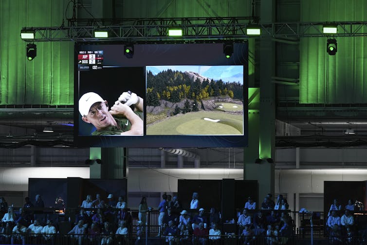 A large screen displaying a virtual gold green and a close up on a man taking a shot hangs above a crowd of people
