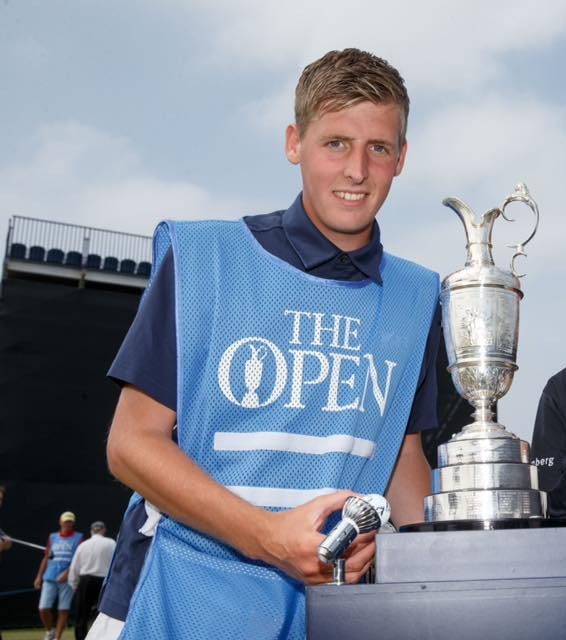 A young man in a "The Open" vest and an older man with a golf club stand beside the Claret Jug.