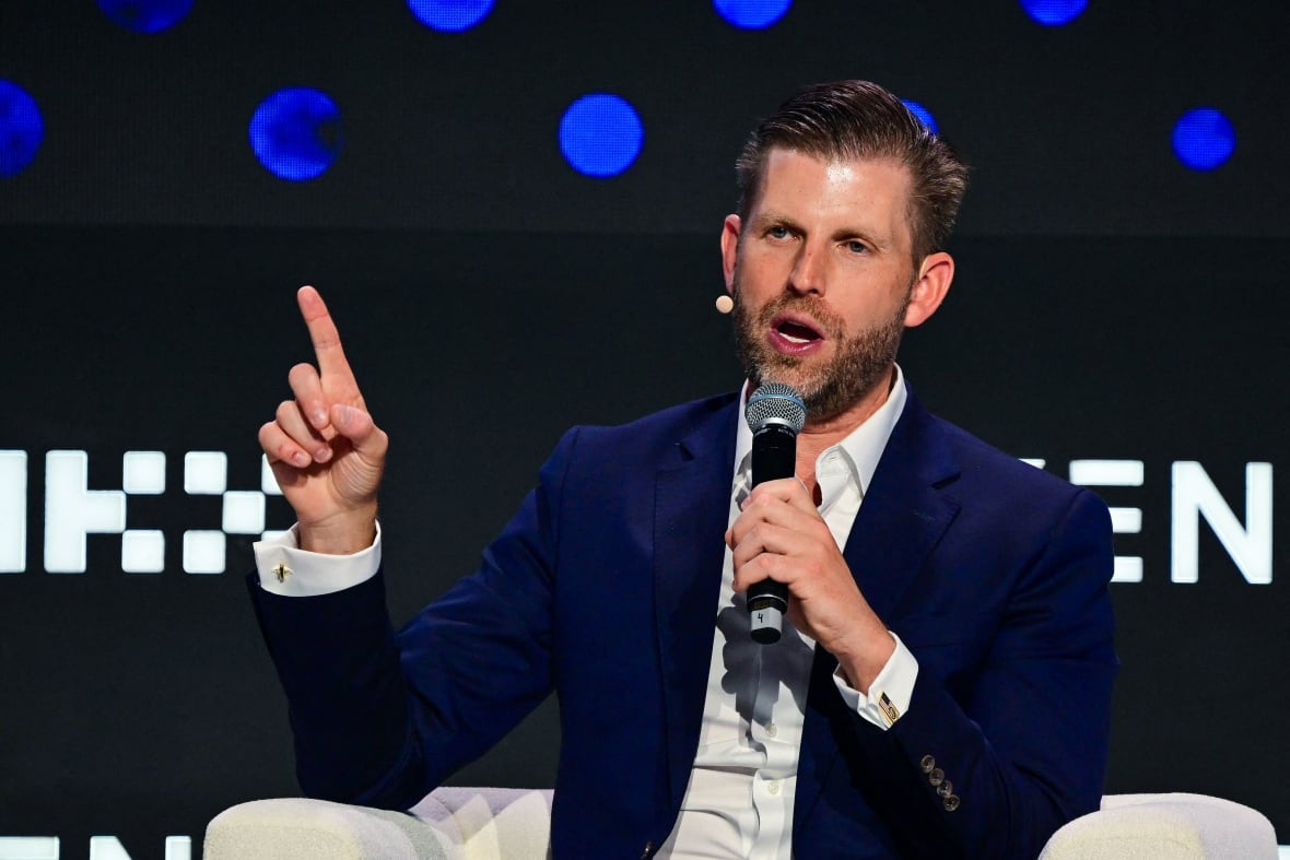 A light-haired bearded man wearing a blazer and a collared shirt is shown seated and speaking into a microphone.