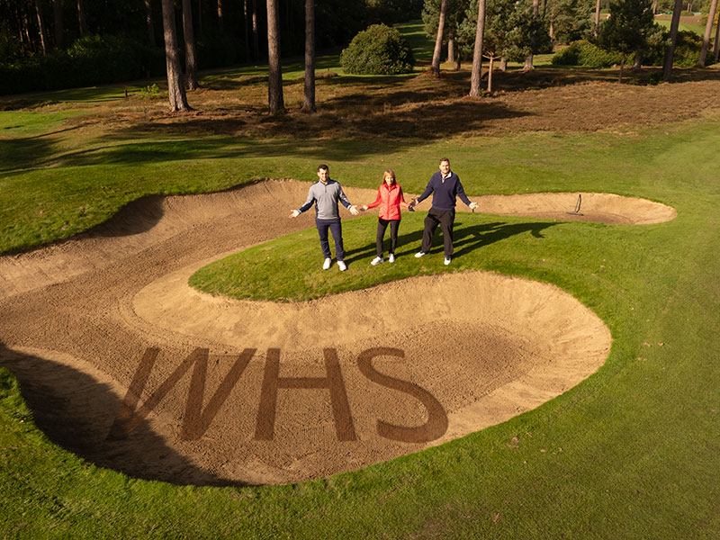 Three golfers shrugging near a bunker with WHS written in the sand