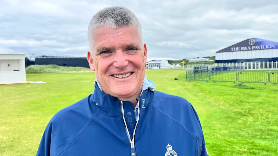 A man with short grey hair wears a blue wet top with a white zip. Behind him blue fencing surrounding a building which say 'The R&A Pavilion' on it. He stands on a patch of green grass.