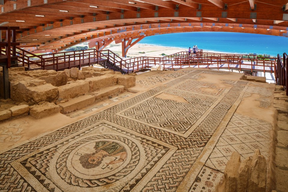 Ancient mosaic floor with geometric and figurative designs, under a modern wooden shelter overlooking the sea.