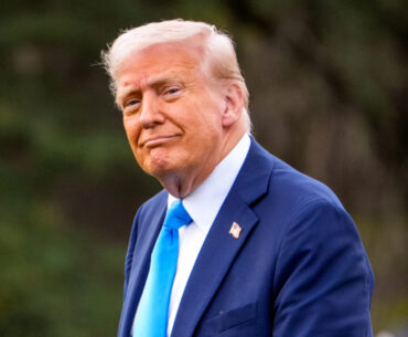 Donald Trump in a blue suit and blue tie, standing outdoors with a slight smile.