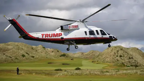 Getty Images A large white, black and red liveried helicopter with TRUMP branding on the fuselage is coming in to land on the fairway at one of the links courses. Trump, in a dark blue suit, can be seen on the edge of frame, standing waiting for it to land. The sky is grey and filled with clouds. 