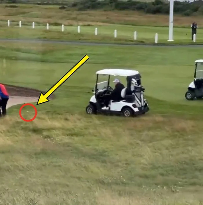 Two golf carts on a green course; one is stuck on a small hill. A person in blue and red clothing stands nearby