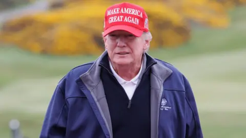 Getty Images Donald Trump in red Make America Great Again golf baseball hat, dark blue Trump Turnberry branded fleece jacket, white polo shirt and blue jumper, is standing in front of a tee at Turnberry. The course can be seen behind him.