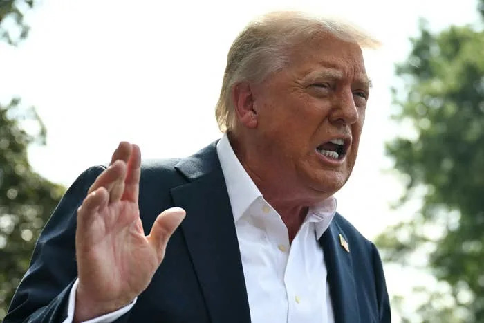 Man in a suit and open-collared shirt speaks outdoors, gesturing with his hand