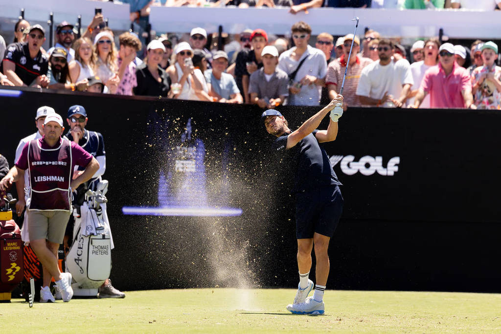 A male golf player swings his putt at LIV Golf. The lawn has been chipped and sprays into the air. The crowd observes from behind him in the stand.