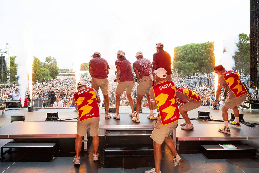 A shot from behind a gold team as they stand on stage in front of a crowd at LIV Golf.