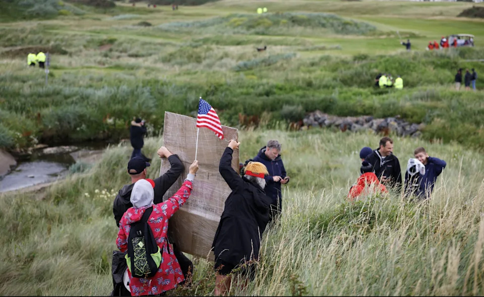 Three of protesters holding an American flag and a placard in the grounds of Turnberry with their backs to the camera. They are standing in a tuft of long grass with a golf course stretching in front of them. The resort's buildings can be seen in the background.