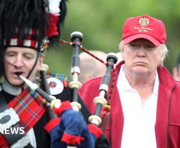 A bagpiper in full military Highland dress with s bearskin hat is playing in the foreground on the left. Behind him and to the right is Donald Trump, in red Trump Golf baseball cap, white shirt and red fleece jacket. He is staring intently at the piper, looking very serious. There are others in golf gear in the background, just out of focus and slightly obscured by the President.