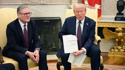 Getty Images Keir Starmer and Donald Trump sitting together in the Oval Office. President Trump is holding a signed invitation from the King. The two men are dressed almost identically in dark blue suits, white shirts and burgundy ties. 
