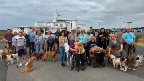 LDRS About 30 people, many with dogs, gather on Birkdale Common to protest plans to allow the R&A to take over a section of the public land ahead of the 2026 Open golf championships there. Three people in wheelchairs sit in the foreground, in front of the standing protesters. 