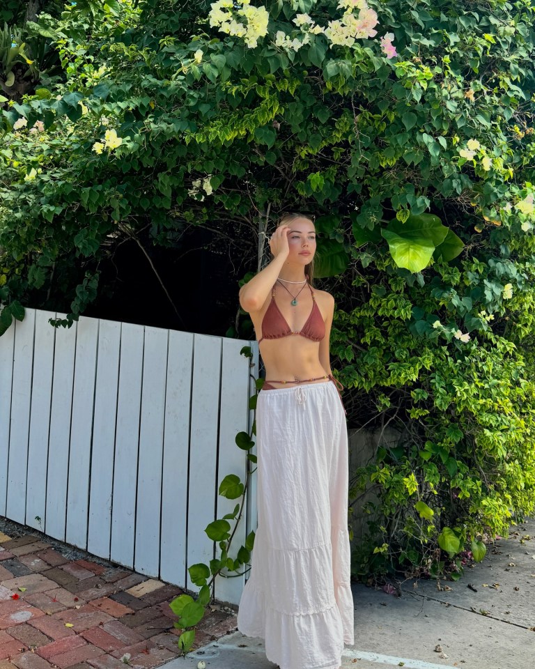 Woman in a brown bikini top and long white skirt standing in front of a white fence and lush green foliage.