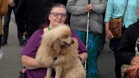 LDRS Chantelle Rea-Bradley, who is sitting on a mobility aid and holding a poodle on her lap. She is seated in front of a group of other protesters. 
