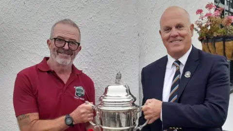 Contributed Stuart Hagger smiles at the camera as he is handed a large silver trophy by another man. Stuart has short grey hair and a grey beard. He wears a red polo T-shirt, glasses and a watch on his wrist. The other man is bald and wears a navy suit with a white shirt and navy tie with some light blue and red stripes on it. 