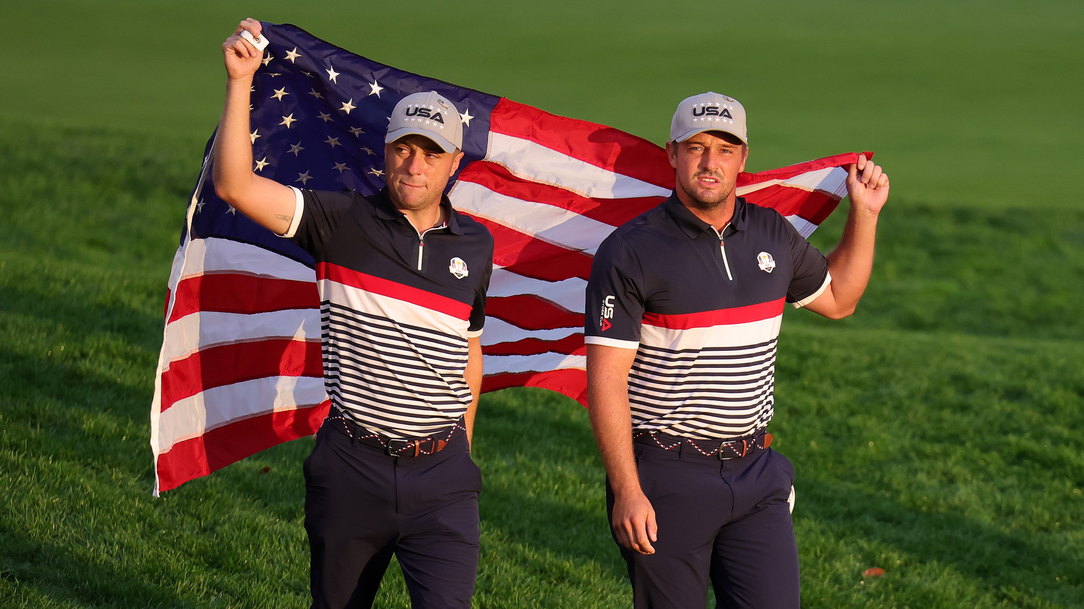 Justin Thomas and Bryson DeChambeau of Team USA carrying an American flag while walking on a golf course.