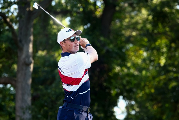 Ben Griffin, in red, white and blue Team USA golf gear, a white cap and sunglasses, hits a drive at the Ryder Cup.