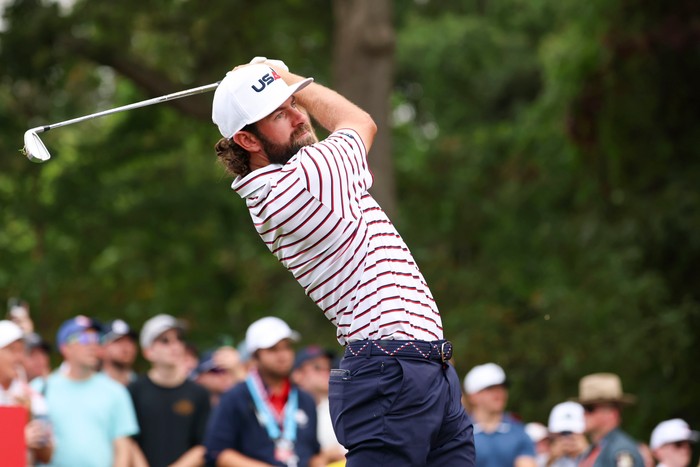 Cameron Young, in white, red and blue striped Team USA golf gear and a white cap, hits a golf shot at the Ryder Cup 2025.