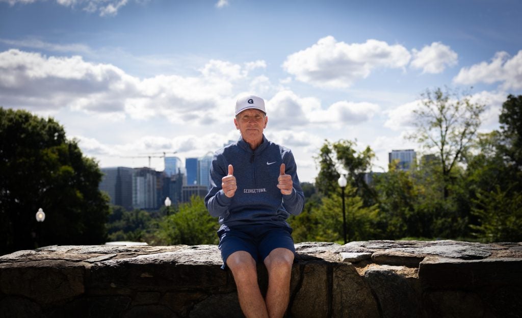 An older Caucasian man in a blue GU quarter zip and a white GU hat with a thumbs up sitting on a ledge on a sunny day