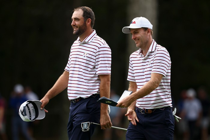 Russell Henley, in white, red and blue striped Team USA golf gear and a white cap, smiles as he walks next to Scottie Scheffler.