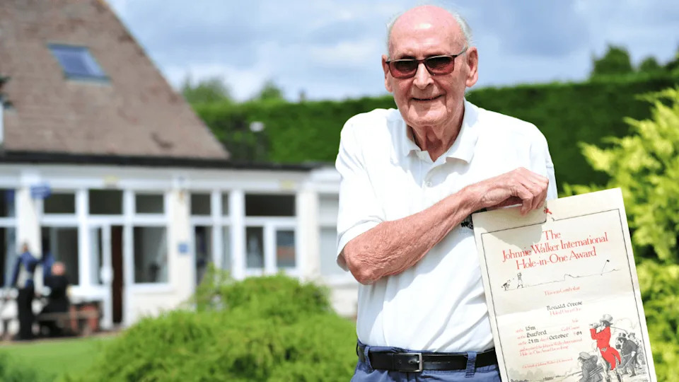 Ron Creese in golfing clothing and holding a 'hole in one' certificate, whilst standing in front of Oxford Golf Club pavilion.