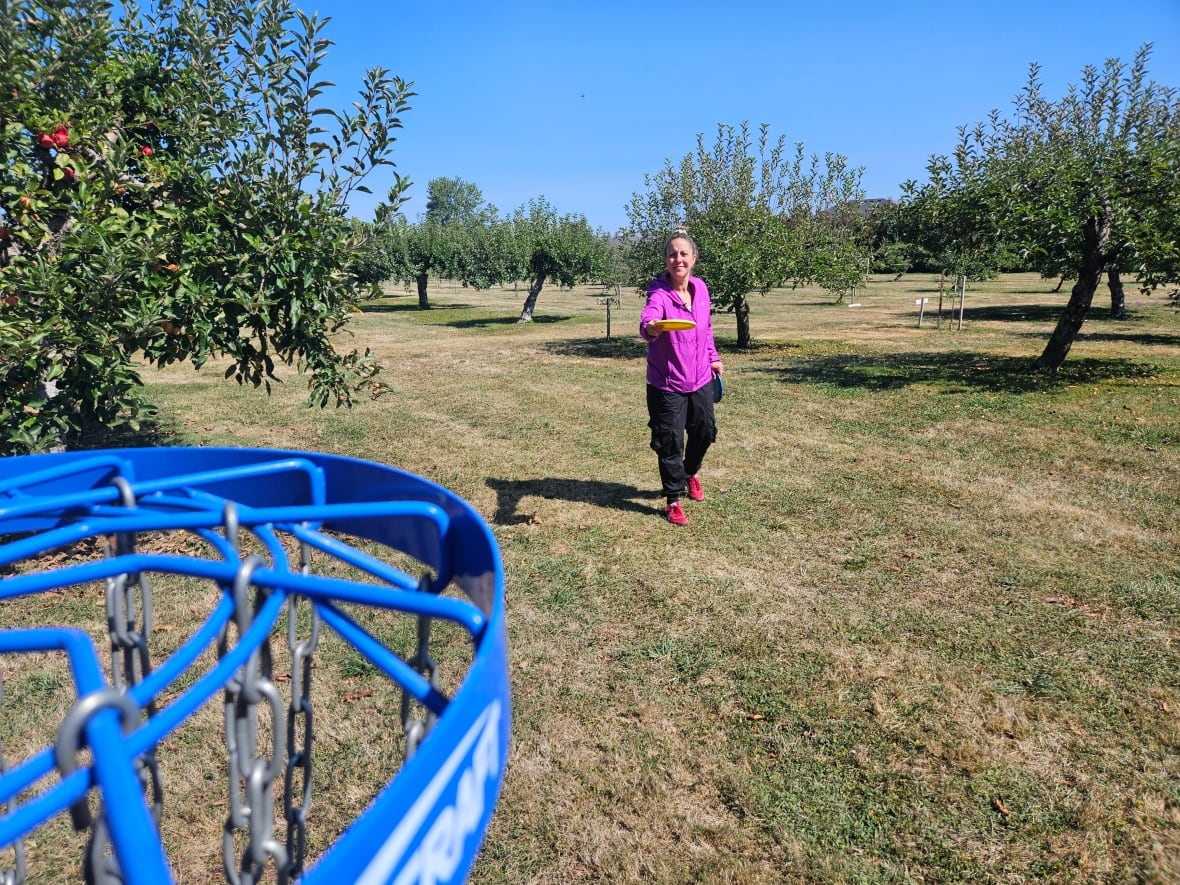 A woman standing in an orchard, throwing a frisbee toward a disc golf target in the foreground. 