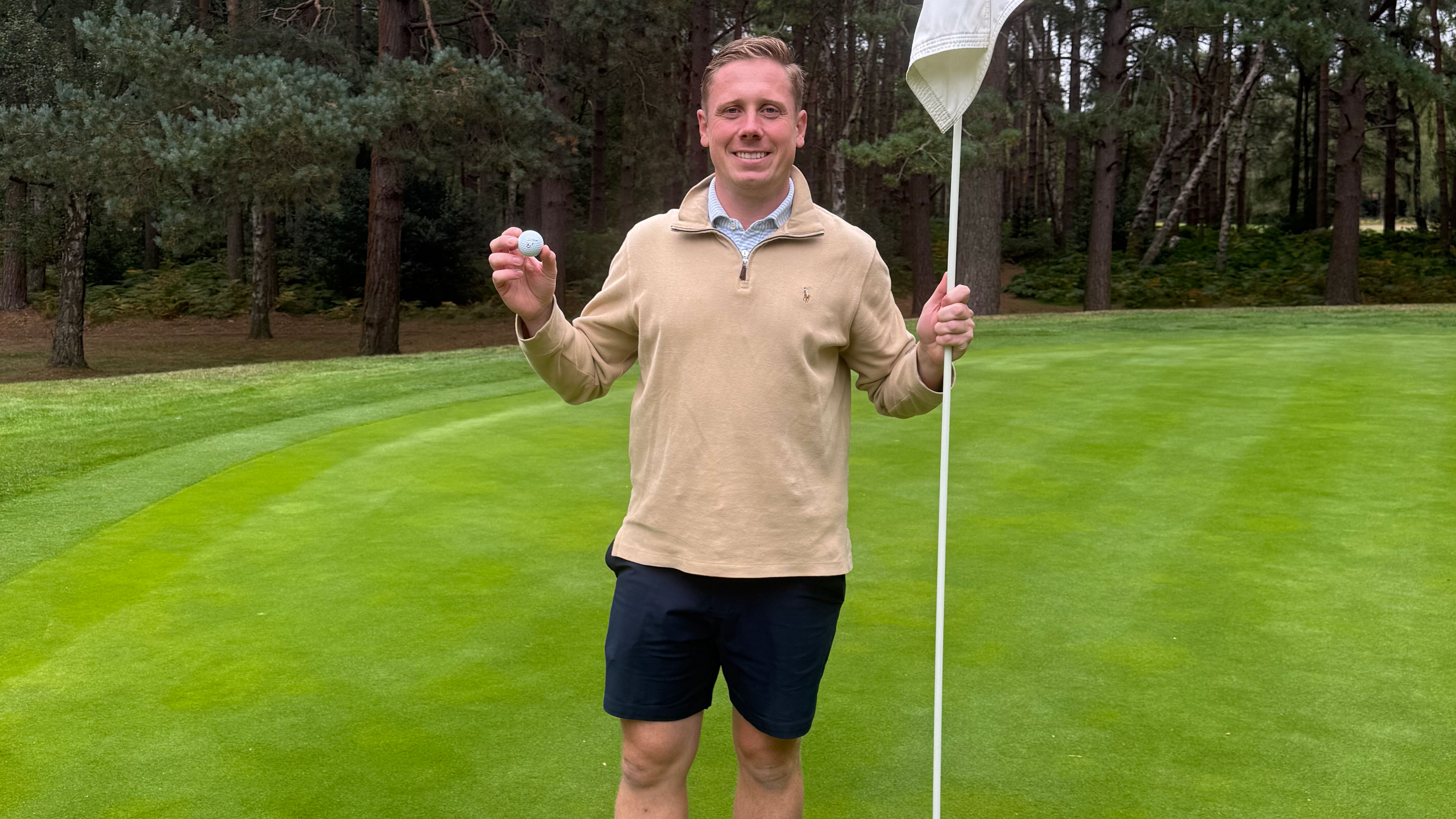 Pete Sawyer holding up his golf ball next to the flag of a par-3 hole after making his third hole-in-one in three months at Woburn Golf Club