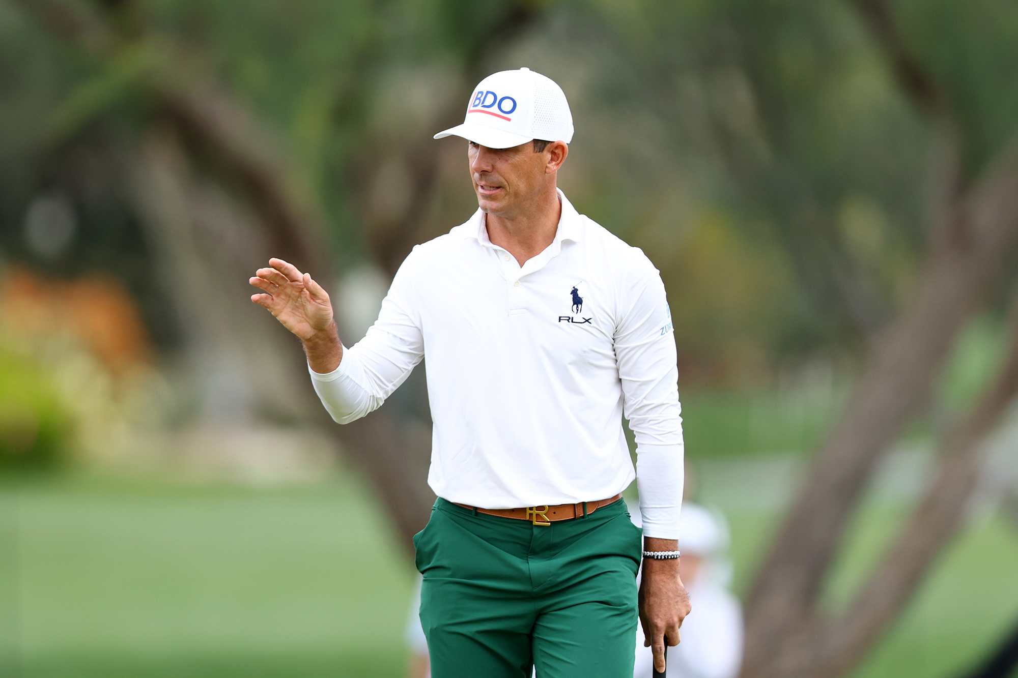 Billy Horschel in a white long sleeved polo shirt and green trousers, holding his hand up to thank the crowd after making a putt at a PGA Tour event